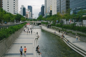 A modern urban setting with a stream running through the center, flanked by paved walkways. People are walking, sitting, and relaxing along the water's edge. Tall buildings and lush green trees line both sides of the stream, creating a blend of natural and constructed elements.