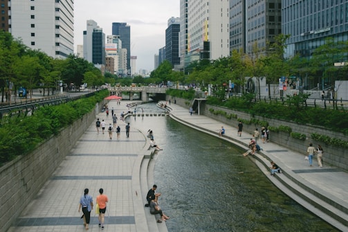 A modern urban setting with a stream running through the center, flanked by paved walkways. People are walking, sitting, and relaxing along the water's edge. Tall buildings and lush green trees line both sides of the stream, creating a blend of natural and constructed elements.
