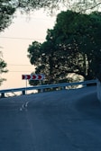 A curved road with a guardrail and directional road signs featuring red and white arrows pointing left. The road is surrounded by lush green trees, and there are utility lines above. The atmosphere seems peaceful, with soft lighting suggesting early morning or late afternoon.