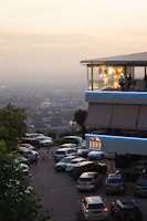A modern restaurant or building with an upper level featuring large windows and decorative yellow lights, set against a hazy cityscape backdrop during sunset. The foreground shows a parking lot with multiple cars and a few people walking nearby. A sign indicates it's a karaoke venue.