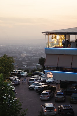 A modern restaurant or building with an upper level featuring large windows and decorative yellow lights, set against a hazy cityscape backdrop during sunset. The foreground shows a parking lot with multiple cars and a few people walking nearby. A sign indicates it's a karaoke venue.