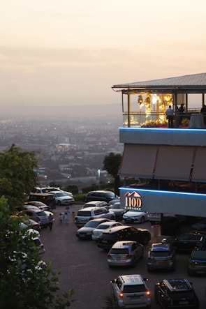 A modern restaurant or building with an upper level featuring large windows and decorative yellow lights, set against a hazy cityscape backdrop during sunset. The foreground shows a parking lot with multiple cars and a few people walking nearby. A sign indicates it's a karaoke venue.