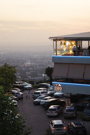 A modern restaurant or building with an upper level featuring large windows and decorative yellow lights, set against a hazy cityscape backdrop during sunset. The foreground shows a parking lot with multiple cars and a few people walking nearby. A sign indicates it's a karaoke venue.