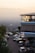 A modern restaurant or building with an upper level featuring large windows and decorative yellow lights, set against a hazy cityscape backdrop during sunset. The foreground shows a parking lot with multiple cars and a few people walking nearby. A sign indicates it's a karaoke venue.