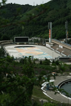 A large, outdoor sports facility nestled in a lush, green hilly area. The complex features an empty skating rink or small stadium surrounded by tiered seating and a wide screen at one end. The stands are marked with advertising banners, and a tall sign with 'JUSAN' is prominently displayed. The foreground has blurred foliage, and there are other structures and pathways nearby.