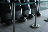 A row of black protective shields are lined up against a wall, each with a matching black helmet placed on the floor. The scene is enclosed by metal stanchions with green tape, and the area appears to be indoors, possibly within a large building or facility.
