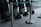 A row of black protective shields are lined up against a wall, each with a matching black helmet placed on the floor. The scene is enclosed by metal stanchions with green tape, and the area appears to be indoors, possibly within a large building or facility.