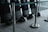 A row of black protective shields are lined up against a wall, each with a matching black helmet placed on the floor. The scene is enclosed by metal stanchions with green tape, and the area appears to be indoors, possibly within a large building or facility.