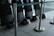 A row of black protective shields are lined up against a wall, each with a matching black helmet placed on the floor. The scene is enclosed by metal stanchions with green tape, and the area appears to be indoors, possibly within a large building or facility.