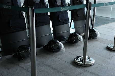 A row of black protective shields are lined up against a wall, each with a matching black helmet placed on the floor. The scene is enclosed by metal stanchions with green tape, and the area appears to be indoors, possibly within a large building or facility.