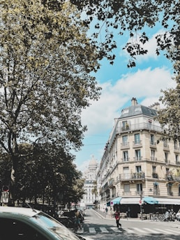 A picturesque city street scene featuring a classic Parisian building with wrought iron balconies and a street-level awning. The street is lined with trees that are lush and green, providing a natural canopy. In the background, the iconic dome of a large church or cathedral is visible. Pedestrians and bicycles are on the street, creating a lively urban atmosphere.