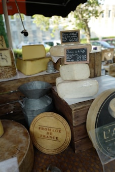 A variety of cheeses displayed on a market stall, including blocks and rounds with small chalkboard signs indicating their names and prices. The cheeses are arranged on wooden crates with a 'Fromage de France' wooden circular sign. There are also metallic containers nearby, and trees can be seen in the background.