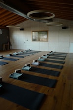 A serene and organized yoga studio with wooden flooring, arranged with several yoga mats evenly spaced across the room. Each mat is equipped with a folded towel and yoga block. The room features a high ceiling with circular light fixtures and a large window on one side, allowing natural light to illuminate the space.