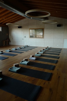 A serene and organized yoga studio with wooden flooring, arranged with several yoga mats evenly spaced across the room. Each mat is equipped with a folded towel and yoga block. The room features a high ceiling with circular light fixtures and a large window on one side, allowing natural light to illuminate the space.