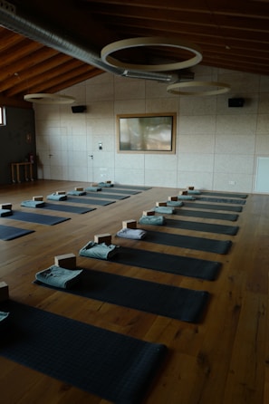 A serene and organized yoga studio with wooden flooring, arranged with several yoga mats evenly spaced across the room. Each mat is equipped with a folded towel and yoga block. The room features a high ceiling with circular light fixtures and a large window on one side, allowing natural light to illuminate the space.