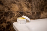 A jar of bio face and body ointment with a partially open lid is sitting on a white ceramic surface. The background features a textured brown marble wall.