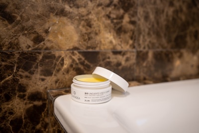 A jar of bio face and body ointment with a partially open lid is sitting on a white ceramic surface. The background features a textured brown marble wall.