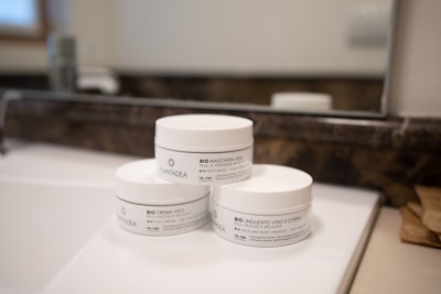 Three white jars of skincare products are arranged on a bathroom counter in front of a mirror. The jars have black text and are labeled as face and body creams with natural ingredients. The background features a faucet and a blurred reflection in the mirror.