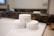 Three white jars of skincare products are arranged on a bathroom counter in front of a mirror. The jars have black text and are labeled as face and body creams with natural ingredients. The background features a faucet and a blurred reflection in the mirror.