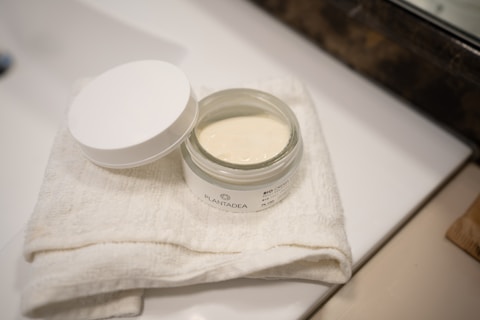 An open jar of cream sits on a folded white towel. The jar has a white lid placed beside it, and the cream inside appears smooth and creamy. The label on the jar displays the brand name 'PLANTADEA'. The setting appears to be a bathroom counter, with a mirror and additional counter space partially visible.