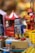 A collection of vintage-style tin toys, including a grey and blue robot with red feet and a painted control panel. In the background, there is a red and white carousel and another toy resembling a fireman. The scene is colorful, with various toys displayed on a shelf.