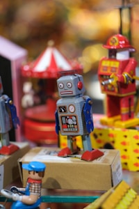 A collection of vintage-style tin toys, including a grey and blue robot with red feet and a painted control panel. In the background, there is a red and white carousel and another toy resembling a fireman. The scene is colorful, with various toys displayed on a shelf.