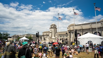 A large crowd of people is gathered in a grassy area in front of an impressive neoclassical building with tall columns. Several American flags are flying on tall poles. People are sitting and standing, some under umbrellas, around a white tent and a prominent monument made of stone statues.