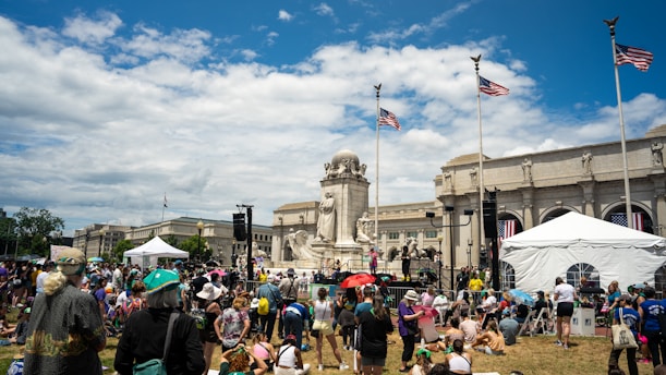 A large crowd of people is gathered in a grassy area in front of an impressive neoclassical building with tall columns. Several American flags are flying on tall poles. People are sitting and standing, some under umbrellas, around a white tent and a prominent monument made of stone statues.