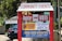A red and white community events sign with multiple smaller signs underneath. The signs advertise various events including voter registration, a car show hosted by the Milford Lions Club, and a Cub Scouts meeting. In the background, there's a black car and greenery.