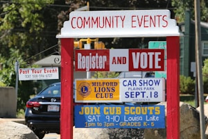 A red and white community events sign with multiple smaller signs underneath. The signs advertise various events including voter registration, a car show hosted by the Milford Lions Club, and a Cub Scouts meeting. In the background, there's a black car and greenery.