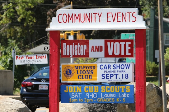 A red and white community events sign with multiple smaller signs underneath. The signs advertise various events including voter registration, a car show hosted by the Milford Lions Club, and a Cub Scouts meeting. In the background, there's a black car and greenery.