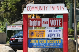 A red and white community events sign with multiple smaller signs underneath. The signs advertise various events including voter registration, a car show hosted by the Milford Lions Club, and a Cub Scouts meeting. In the background, there's a black car and greenery.