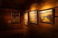 A cozy, warmly lit room with wooden walls features three framed paintings hanging on the wall. The artwork includes colorful nature scenes, floral imagery, and abstract landscapes. The wood paneling adds a rustic charm to the setting.
