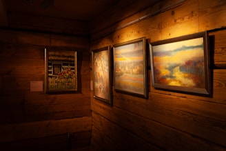 A cozy, warmly lit room with wooden walls features three framed paintings hanging on the wall. The artwork includes colorful nature scenes, floral imagery, and abstract landscapes. The wood paneling adds a rustic charm to the setting.