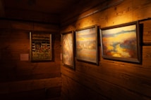 A cozy, warmly lit room with wooden walls features three framed paintings hanging on the wall. The artwork includes colorful nature scenes, floral imagery, and abstract landscapes. The wood paneling adds a rustic charm to the setting.