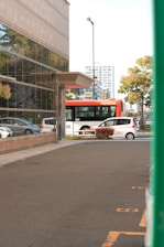 An urban street scene featuring a large glass building on the left, reflecting the surrounding trees and sky. A red bus marked 'IYOTETSU' is partially visible behind the building, and a white car is parked nearby. The street is mostly empty, and a few trees with green foliage are in the background alongside several tall buildings.