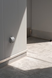 A minimalistic architectural setting with smooth, light gray walls and floor tiles. There's a shadow cast diagonally across the scene creating a contrast between light and shadow. The area appears clean and modern with a small metallic fixture visible on the wall and a hinge on the door.