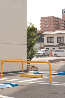 A parking lot features yellow and blue wheel stops, with metal barriers. The area is bordered by residential and commercial buildings, including a multi-story apartment block in the background. A tree is next to a white wall on the left, and there are parked cars and a sign indicating a 'For Rent' notice.