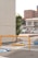 A parking lot features yellow and blue wheel stops, with metal barriers. The area is bordered by residential and commercial buildings, including a multi-story apartment block in the background. A tree is next to a white wall on the left, and there are parked cars and a sign indicating a 'For Rent' notice.
