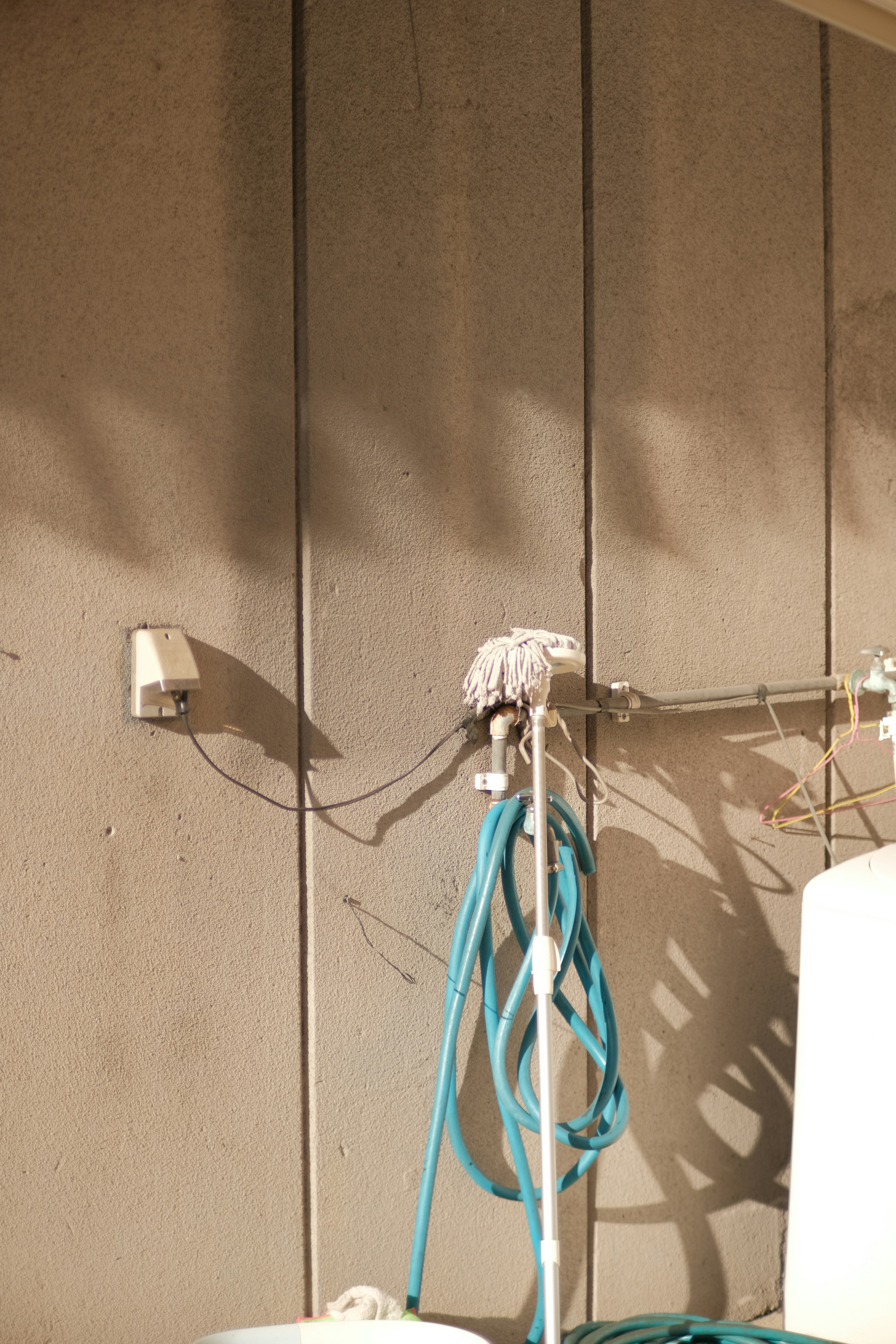 A concrete wall with a mounted rack holding a mop and a coiled blue hose. There is a small electrical box on the wall, and sunlight casts shadows of the objects onto the surface.