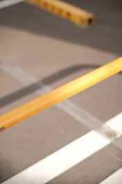 A close-up view of a yellow barrier pole positioned diagonally over asphalt, with part of a parking lot's striped markings visible in the background.