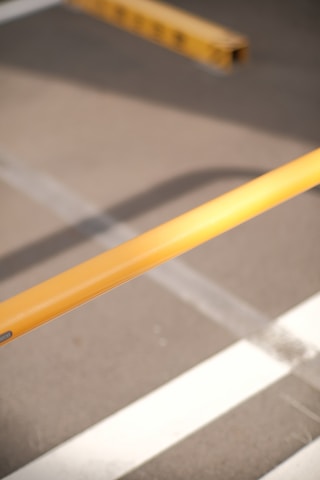 A close-up view of a yellow barrier pole positioned diagonally over asphalt, with part of a parking lot's striped markings visible in the background.