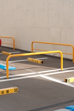 A parking area with yellow metal barriers and painted white lines on the asphalt. Japanese warning signs are positioned near the barriers. The backdrop is a plain concrete wall.