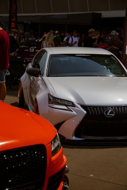 A car show featuring a white Lexus in the foreground and an orange Audi partially visible. The setting is outdoors with many people gathered, some looking at the cars while others engage in conversations. The crowd includes individuals dressed casually in summer attire.