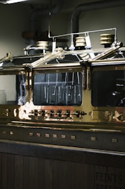 An industrial metallic machine with a polished brass surface, featuring several dials, knobs, and what seem to be pressure gauges on top. The machine is encased in a glass enclosure, reflecting the overhead lights. The environment appears to be a distillery or similar industrial setting.