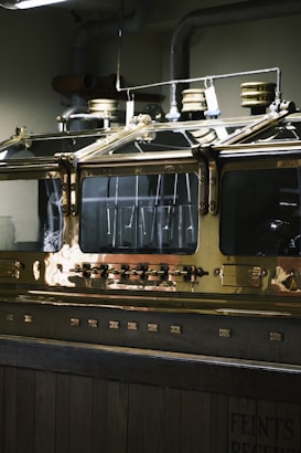 An industrial metallic machine with a polished brass surface, featuring several dials, knobs, and what seem to be pressure gauges on top. The machine is encased in a glass enclosure, reflecting the overhead lights. The environment appears to be a distillery or similar industrial setting.