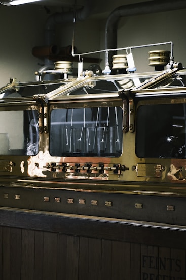 An industrial metallic machine with a polished brass surface, featuring several dials, knobs, and what seem to be pressure gauges on top. The machine is encased in a glass enclosure, reflecting the overhead lights. The environment appears to be a distillery or similar industrial setting.