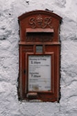 A vintage red postal box embedded in a white textured wall, featuring an emblem and the inscription 'G R' at the top. Below is a slot labeled 'Letters Only' and further down is a notice with collection times for mail from Monday to Saturday.