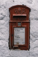 A vintage red postal box embedded in a white textured wall, featuring an emblem and the inscription 'G R' at the top. Below is a slot labeled 'Letters Only' and further down is a notice with collection times for mail from Monday to Saturday.