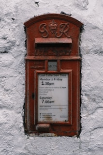 A vintage red postal box embedded in a white textured wall, featuring an emblem and the inscription 'G R' at the top. Below is a slot labeled 'Letters Only' and further down is a notice with collection times for mail from Monday to Saturday.